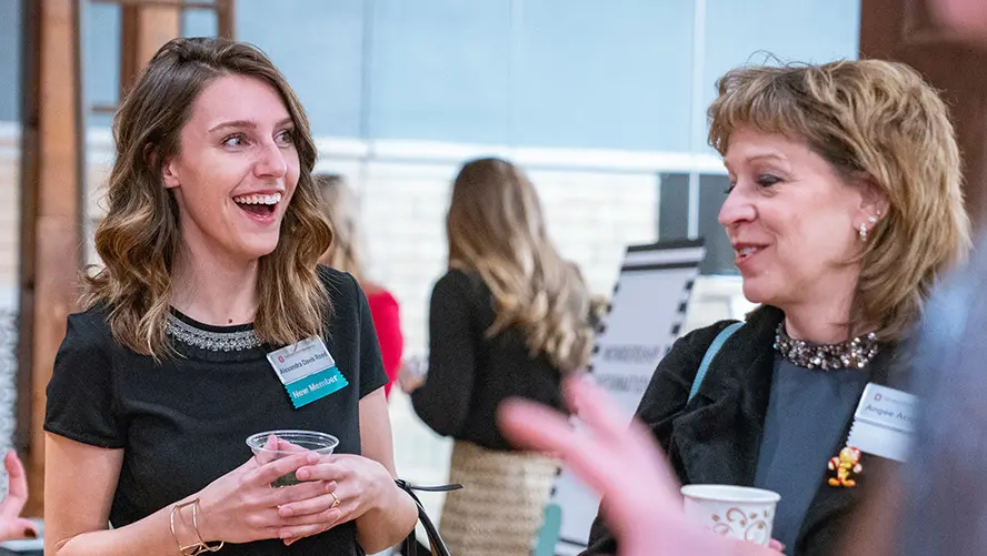 Two women are in a discussion while at an event. They are wearing names tags and holding disposable cups.