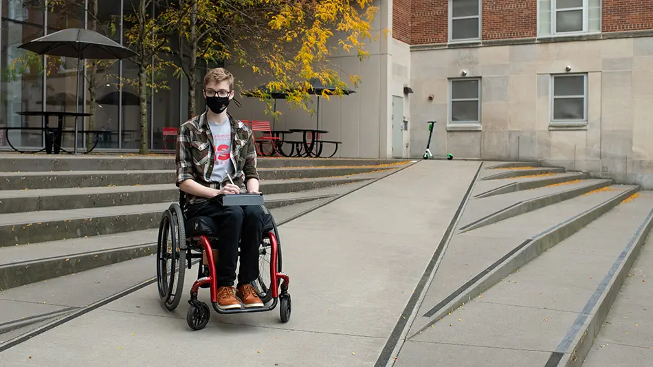 Kayden Gill is seated in his red wheelchair on a ramp that crosses through exterior steps to a building