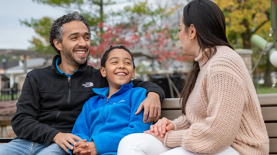 Jorge Ramirez Orrego is sitting on a park bench with his arm around his son, Simon, with his wife, Tatiana Cuellar, sitting next to Simon.