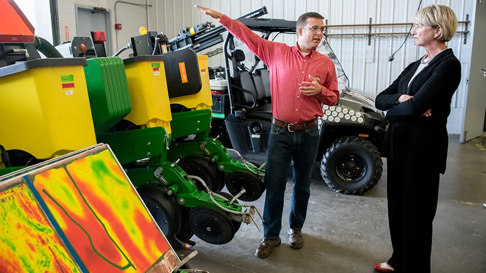 man shows farm equipment as a woman in a suit listens attentively