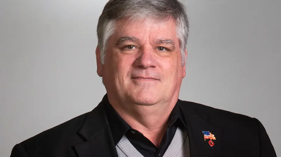 Portrait of Kelley Crooks wearing a dark suit jacket and a united states flag pin on his lapel