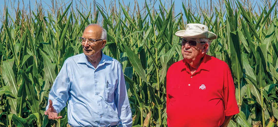 Two men are standing in front of corn stalks. One is wearing a light blue long sleeve button-up and the other is wearing a red polo shirt and a tan hat.