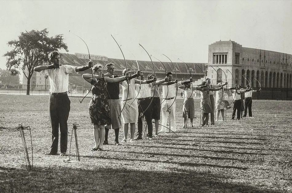 Black and white archival photo of an archery class outside on the lawn. Each student has a bow and is pulling back the string as they prepare to shoot.