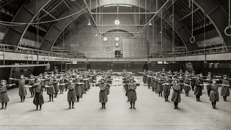 Black and white archival photo of a large group of women lined up in rows with their arms extended straight out from their sides. They are in a large space with a curved roof and a skylight.