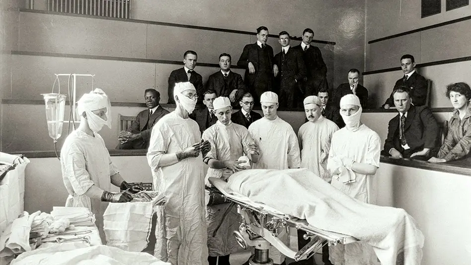 Black and white archival photo of an operating room from 1917. 6 doctors are performing surgery while a group watches from raised benches around the perimeter of the room.