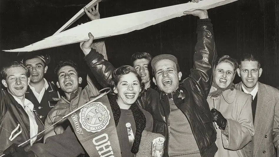 Black and white archival photo of a group of male and female students after a football game. There is a man in the center holding a large shard of wood from the goal post about his head.
