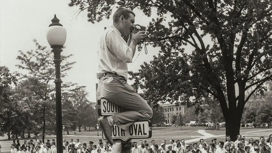 Black and white archival photo of a large gathering of people watching a parade. In the foreground is a man who has climbed up to the top of a street sign holding a camera. 