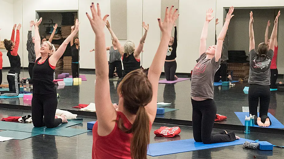 A yoga class in a studio with the back wall made of mirrors. The women in the class are kneeling on their mats with their arms raised straight up next to their ears.