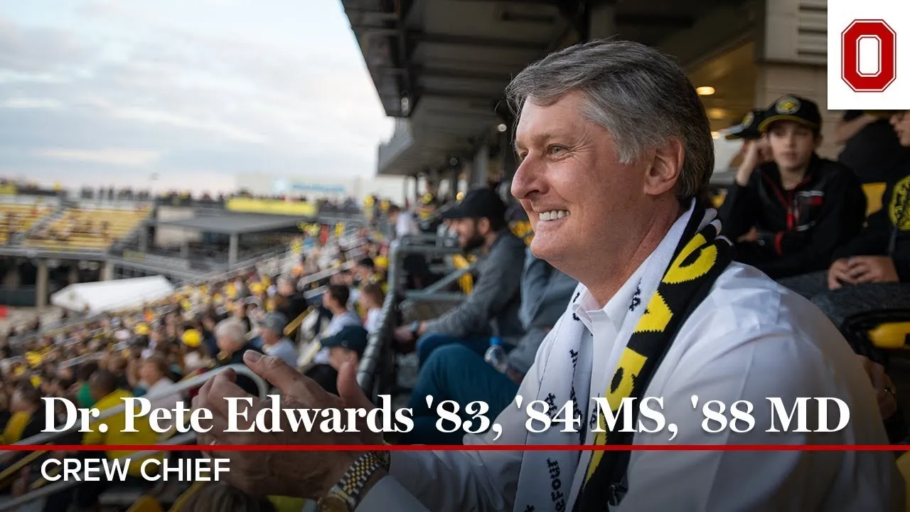 Edwards is clapping while watching the Columbus Crew soccer match inside Mapfre Stadium. He is smiling and wearing a white long sleeve shirt and a black and yellow Crew scarf.