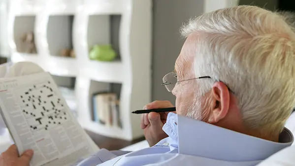 An older man is seated in a chair while doing a crossword puzzle with a pen.