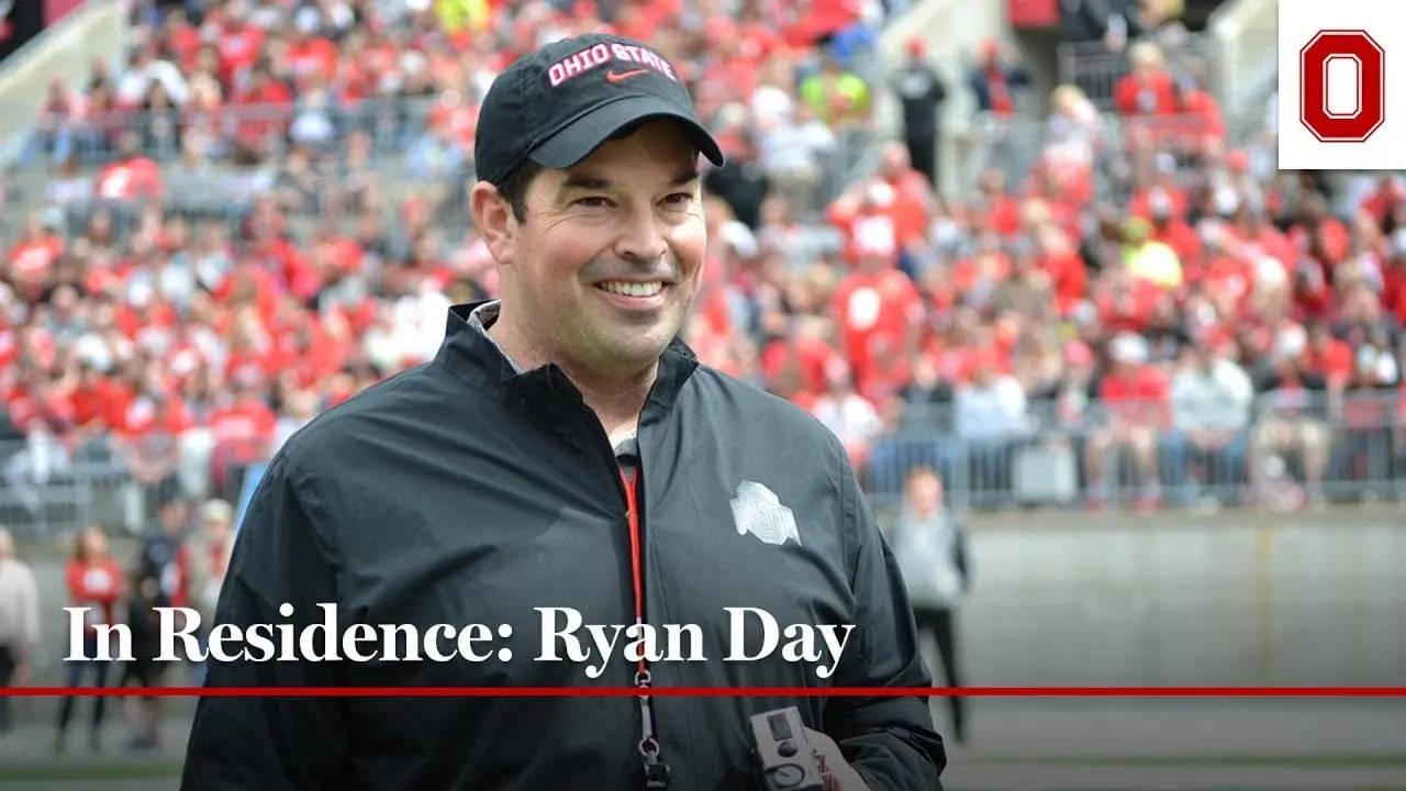 Ryan Day is standing in Ohio Stadium, smiling, with a view of the fans in the background. He is wearing a black jacket and ball cap 