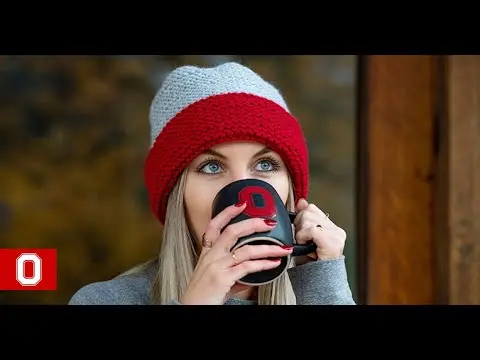 A young woman is wearing a gray and red knit winter hat and drinking from a black and red OSU mug