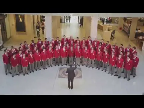 Aerial photo of the Ohio State Men's Glee Club standing in a semi-circle with a conductor standing in the center. The men are wearing red suit jackets and tan pants, while the conductor wears all black.