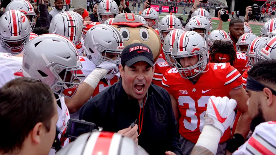 Head coach of the Ohio State football team is standing in the middle of a huddle of players in football uniforms. He is wearing a black long sleeve shirt and a black OSU hat.