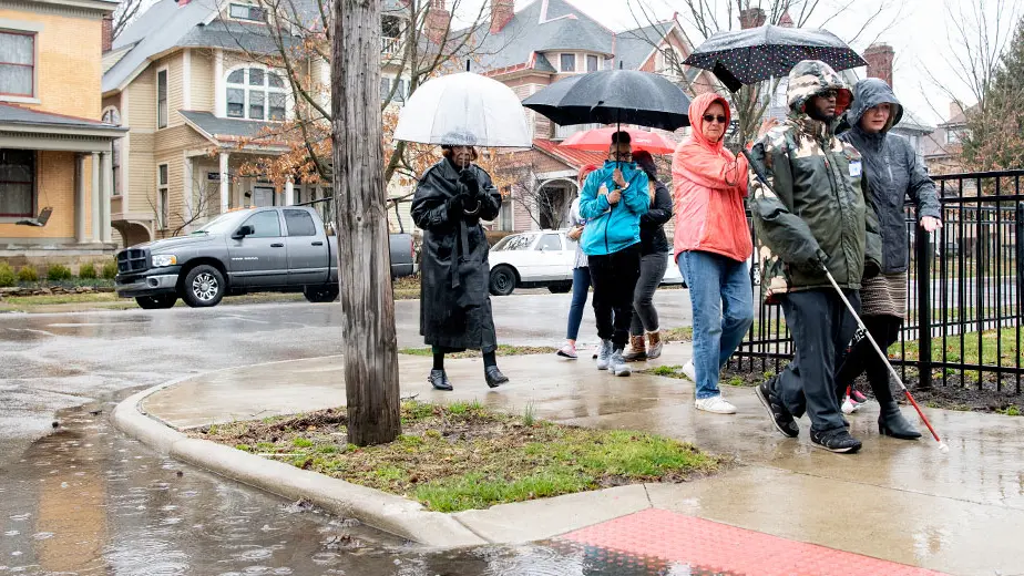 group of people walking in the rain behind a blind man using a cane