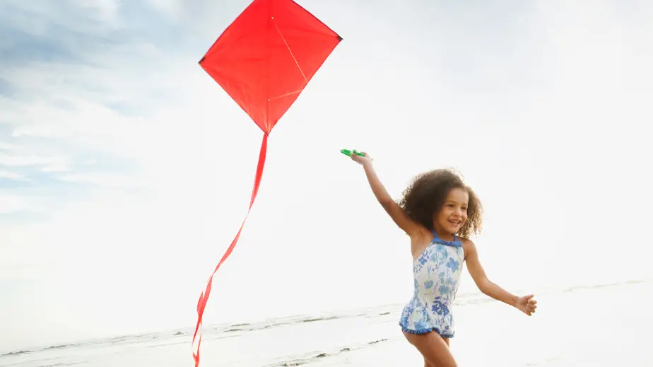 young girl in swimsuit flying a red kite on the beach