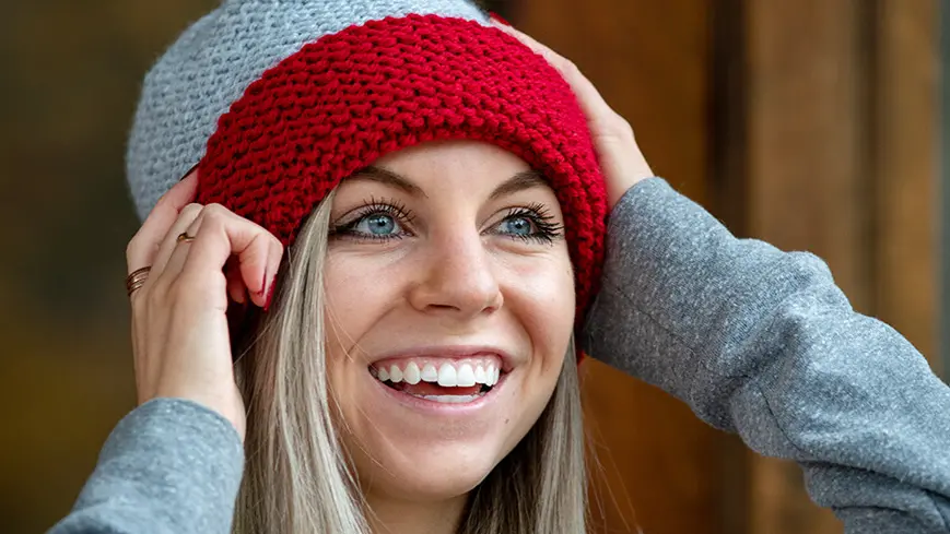 A young woman is wearing a gray and red knit winter hat and smiling