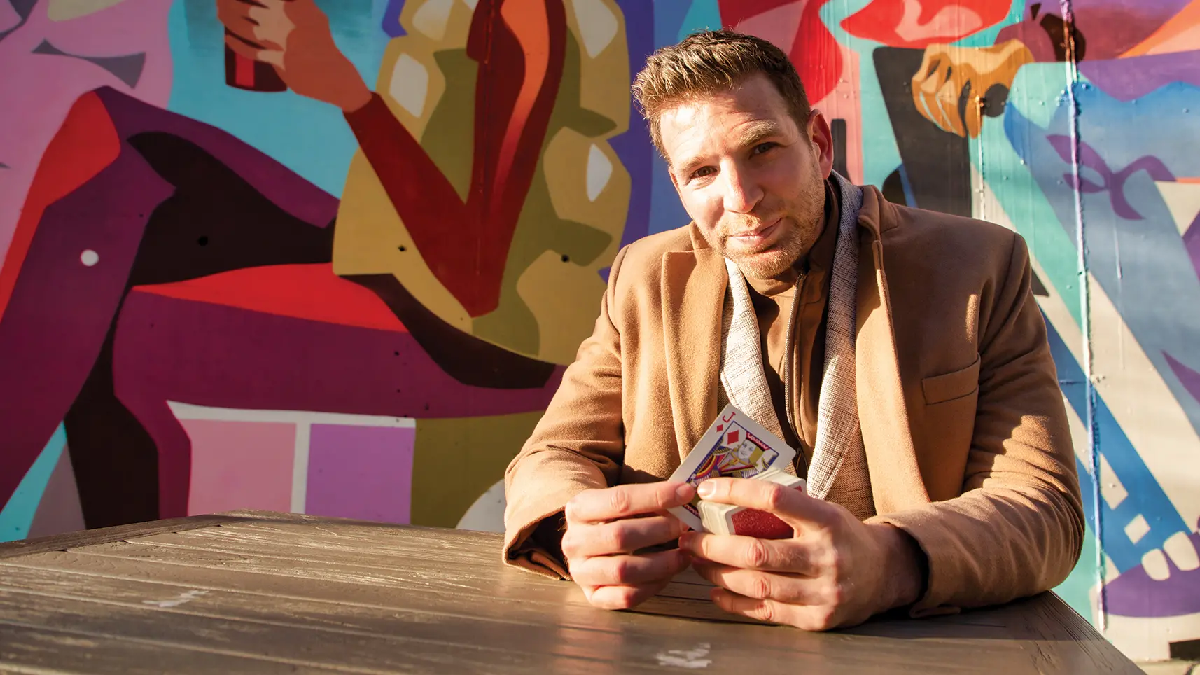 Smiling with his eyes, Joshua Jay holds a jack of diamonds while sitting at an outside mural