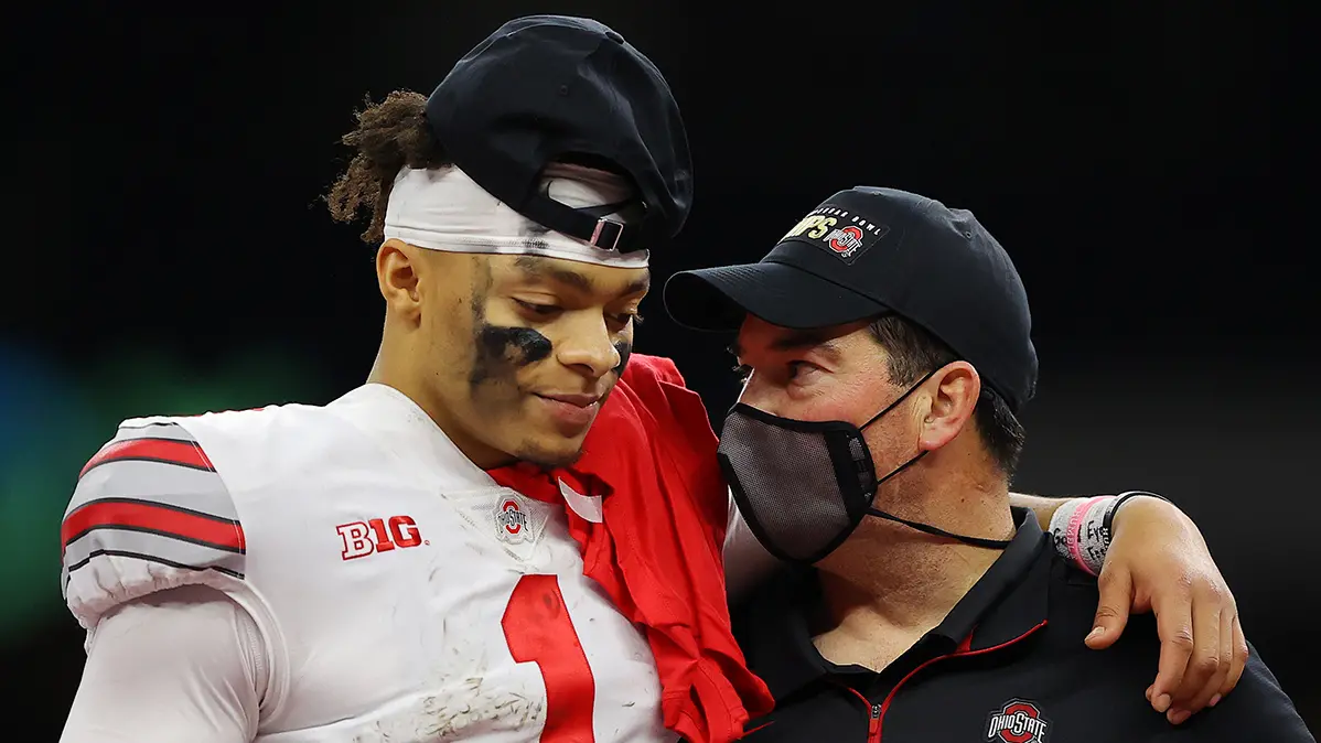 Image of Justin Fields, OSU football player in white jersey with arm around Coach Day, who is wearing a gray cloth face mask and a black Champs hat. White text overlaid says "Ryan Day reflects on 2020 and looks ahead" 