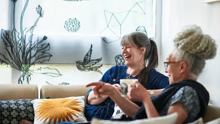 Two woman sit at home laughing, enjoying coffee, and each others company