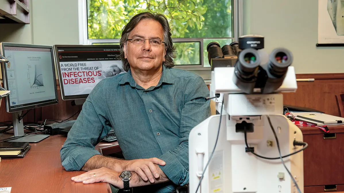 Photo of white man with glasses and blue button up shirt next to a microscope. 