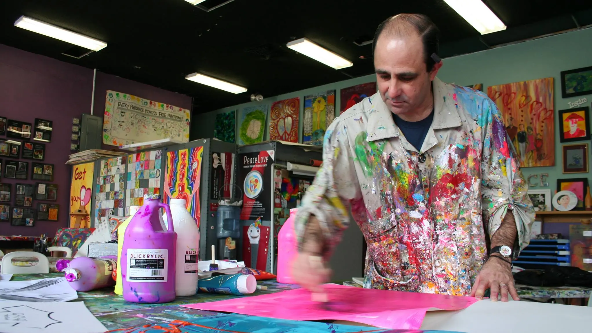 Wearing a paint-splattered smock, a man slightly smiles slightly while painting in his colorful studio.