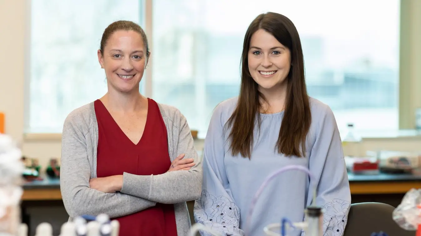 Two women stand next to each other, smiling in a lab setting.