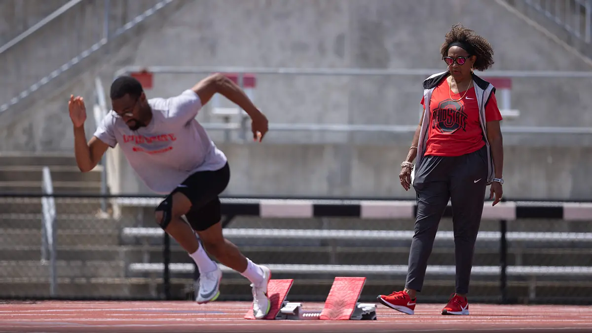 A young black man seems to sprint off the left side of the photo as his coach, standing aside, monitors his form