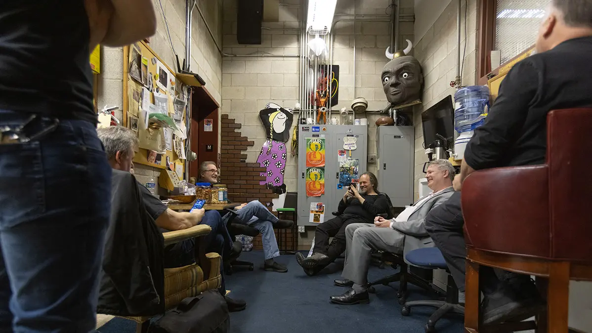 In a narrow room behind a stage, old props line the walls and six men smile while talking and laughing.