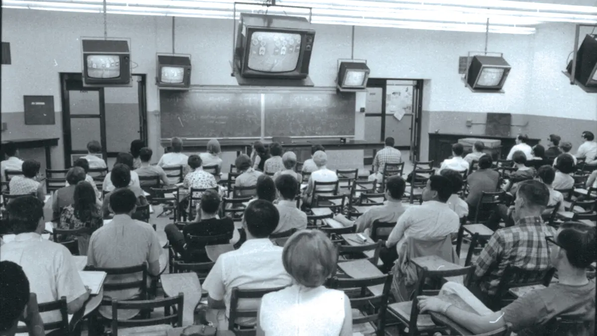 A black and white photo shows a large classroom with straight-backed chairs, old-fashioned TVs mounted near the ceiling and a crowd of students. 