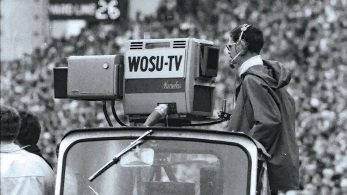 A black and white photo shows an old-style television camera and cameraman in Oho Stadium. 