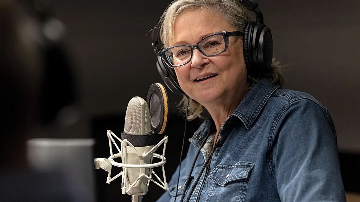 An older white woman wears glasses and headphones as she attentively listens to someone, slightly smiling. A microphone sits in front of her.  