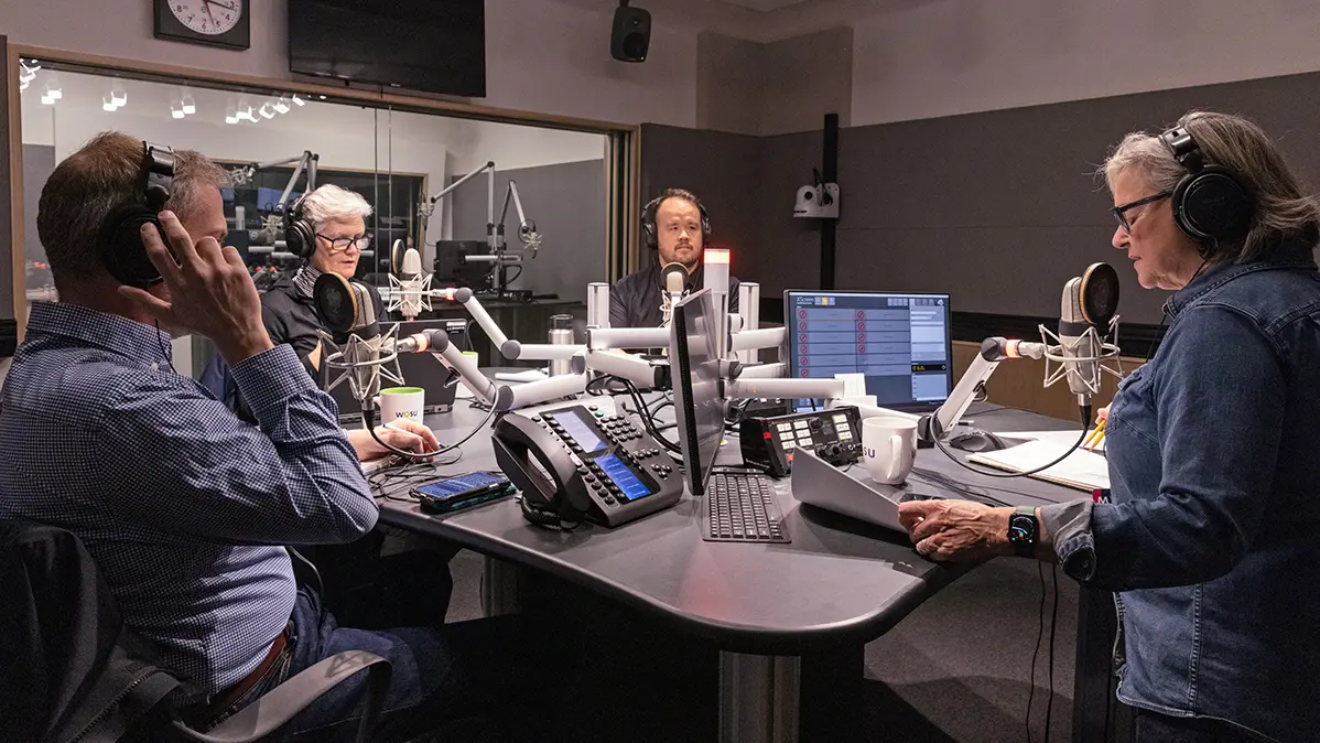 Four people, all wearing headphones, sit at a table loaded with computers and microphones on long, adjustable arms. They’re talking as a radio show is broadcast. 