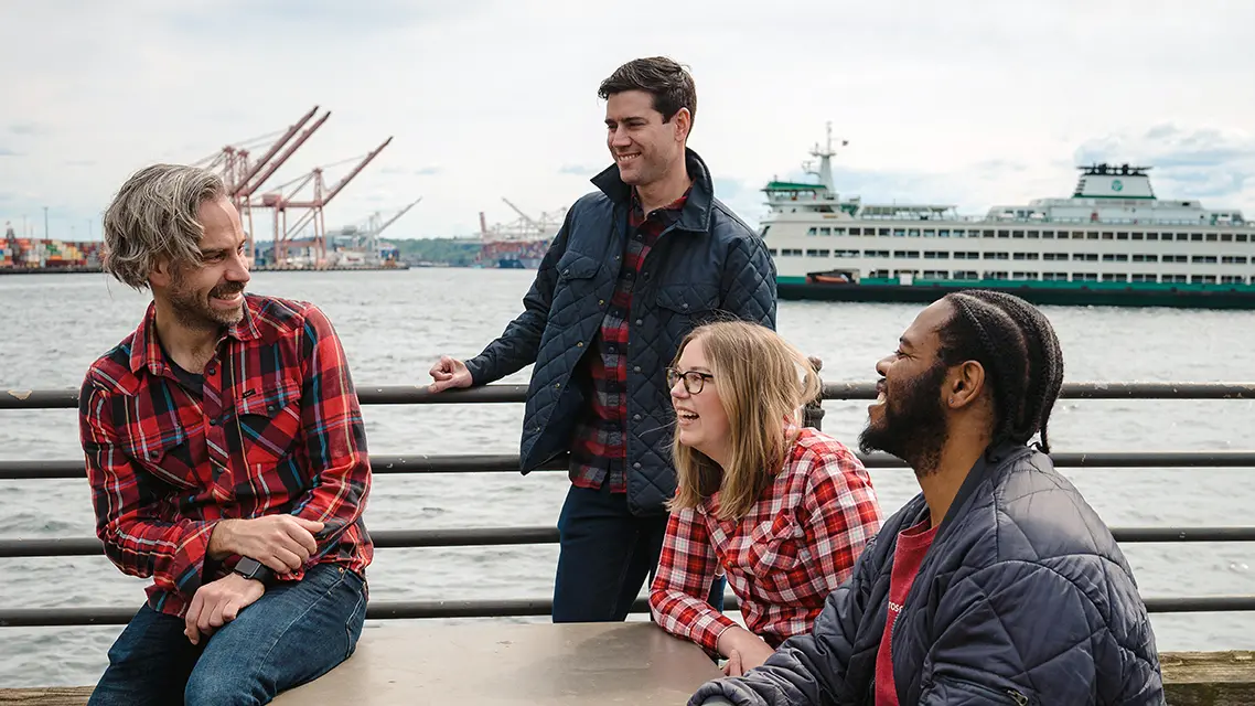 Four people, three wearing red plaid, laugh together around a table on the waterfront. A big ferry and cranes can be seen behind them.