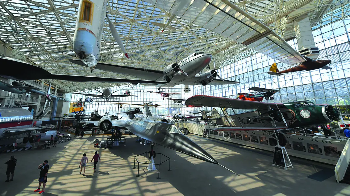Airplanes hanging from the ceiling and parked on the ground look fast even while standing still inside a glass-and-metal-frame hangar. Blue skies can be seen outside and people in the museum look small.