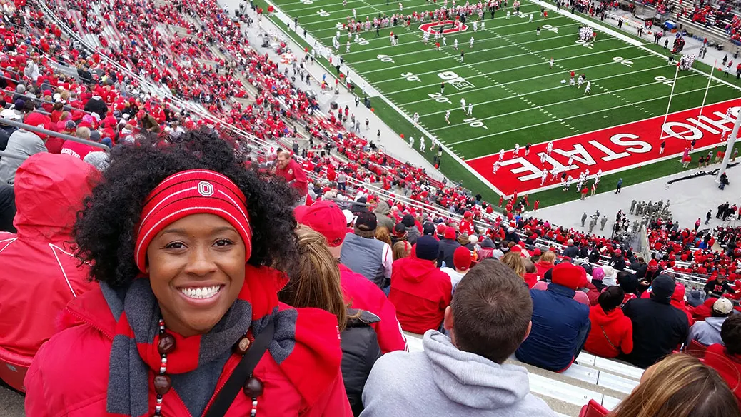 Inside the ’Shoe, a happy woman decked out in Ohio State gear sits high in the stands as football players line the field behind her.