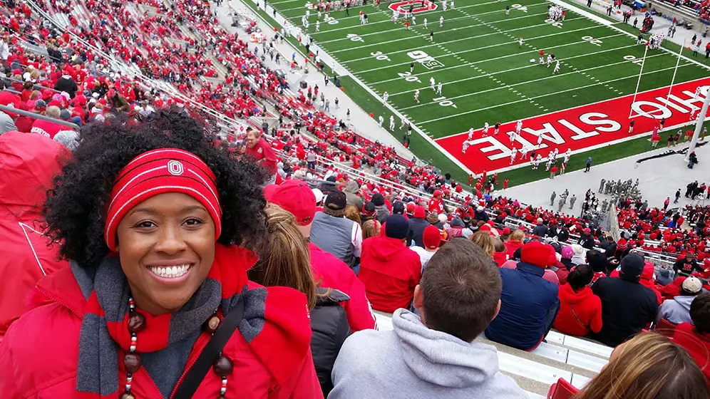 Inside the ’Shoe, a happy woman decked out in Ohio State gear sits high in the stands as football players line the field behind her.