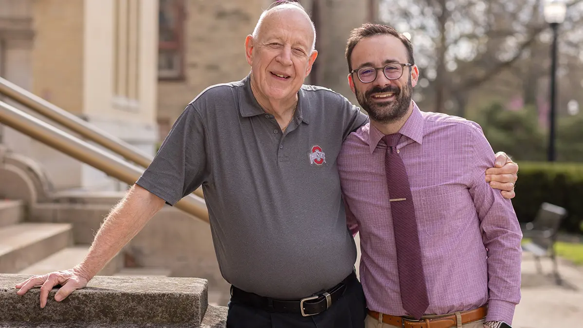 An older man in an Ohio State golf shirt and a younger man wearing a button-up shirt and tie stand side by side with arms around each other. They look relaxed and happy.