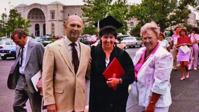 In an old color photograph, a young woman in cap and gown stands between her proud parents. Ohio Stadium can be seen in the background.
