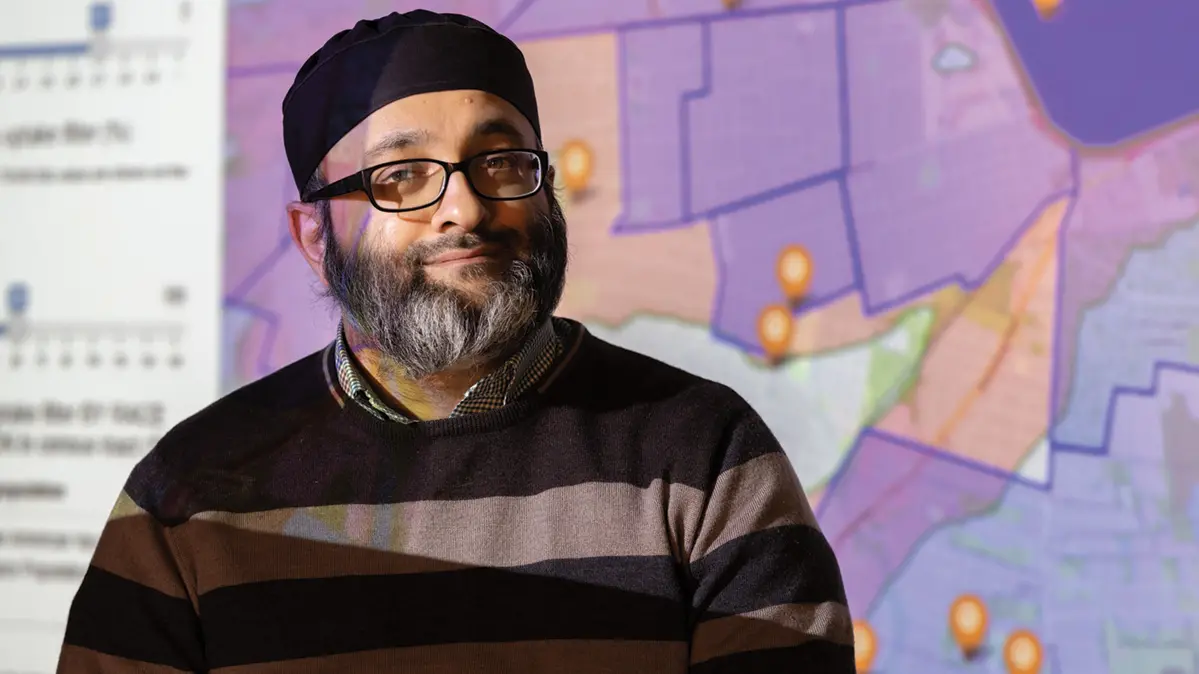A bearded man wearing glasses and a kufi looks directly at the viewer and slightly smiles. A colorful map is projected onto him and the wall behind him. 