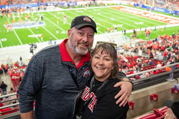 Both smiling, a man has his arm around his wife in the stands at a football game