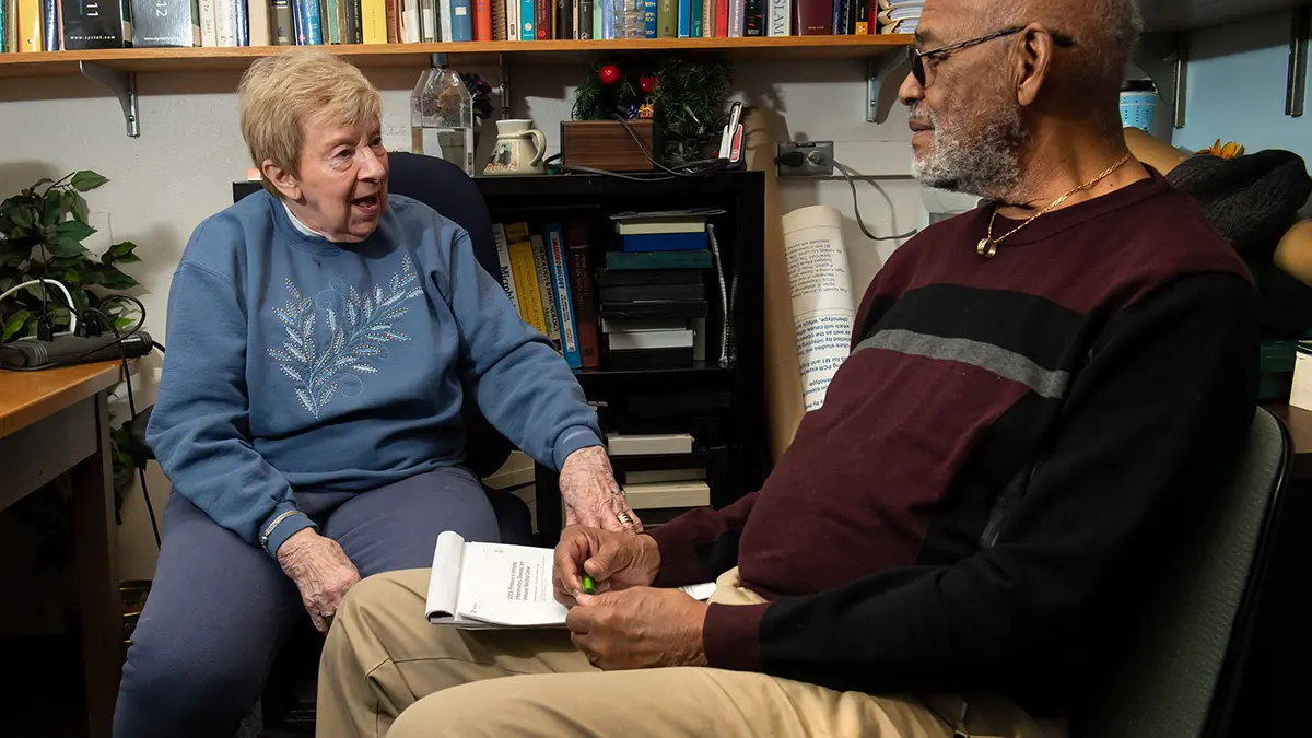 An older white woman in a book-lined professor’s office touches the hand of an older Black man as they reminisce.