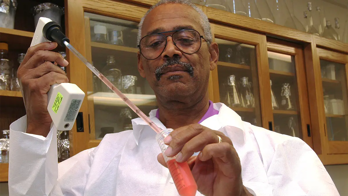 A Black man in a lab coat focuses and he uses scientific instruments to measure a red fluid.