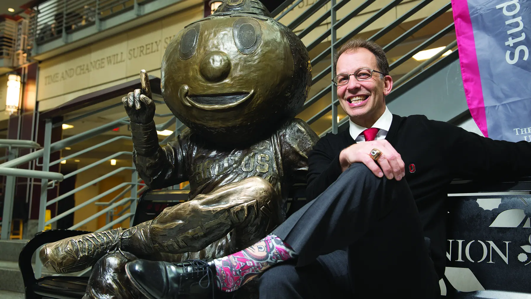 A man smiles as he sits on a bench next to a bronze statue of Brutus. Their leg-crossed poses and smiles are similar.