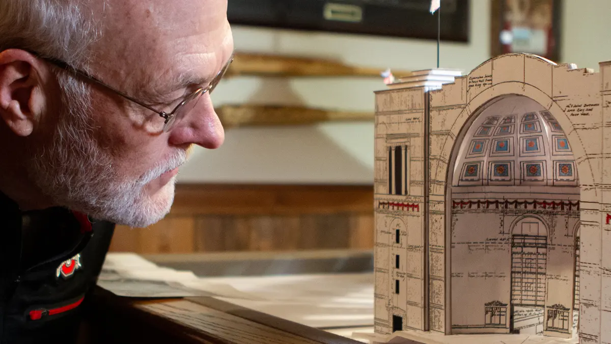 In a basement decked out with Ohio State keepsakes, an older man in glasses takes a close look at the front of the Ohio Stadium model he built, which is slightly taller than his head.