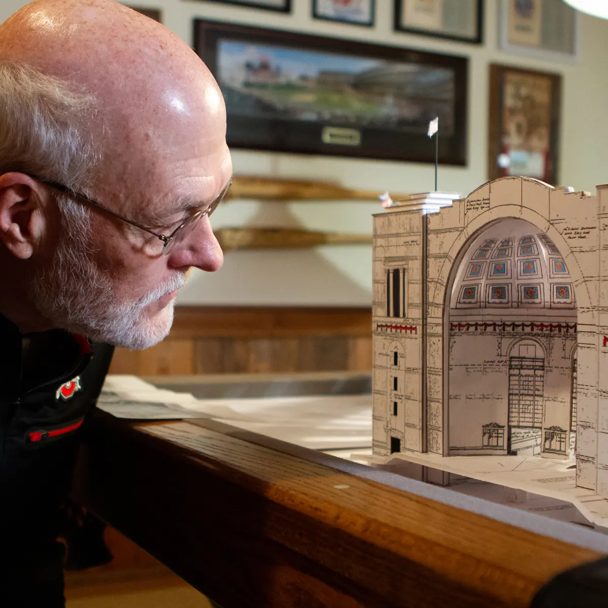 In a basement decked out with Ohio State keepsakes, an older man in glasses takes a close look at the front of the Ohio Stadium model he built, which is slightly taller than his head.