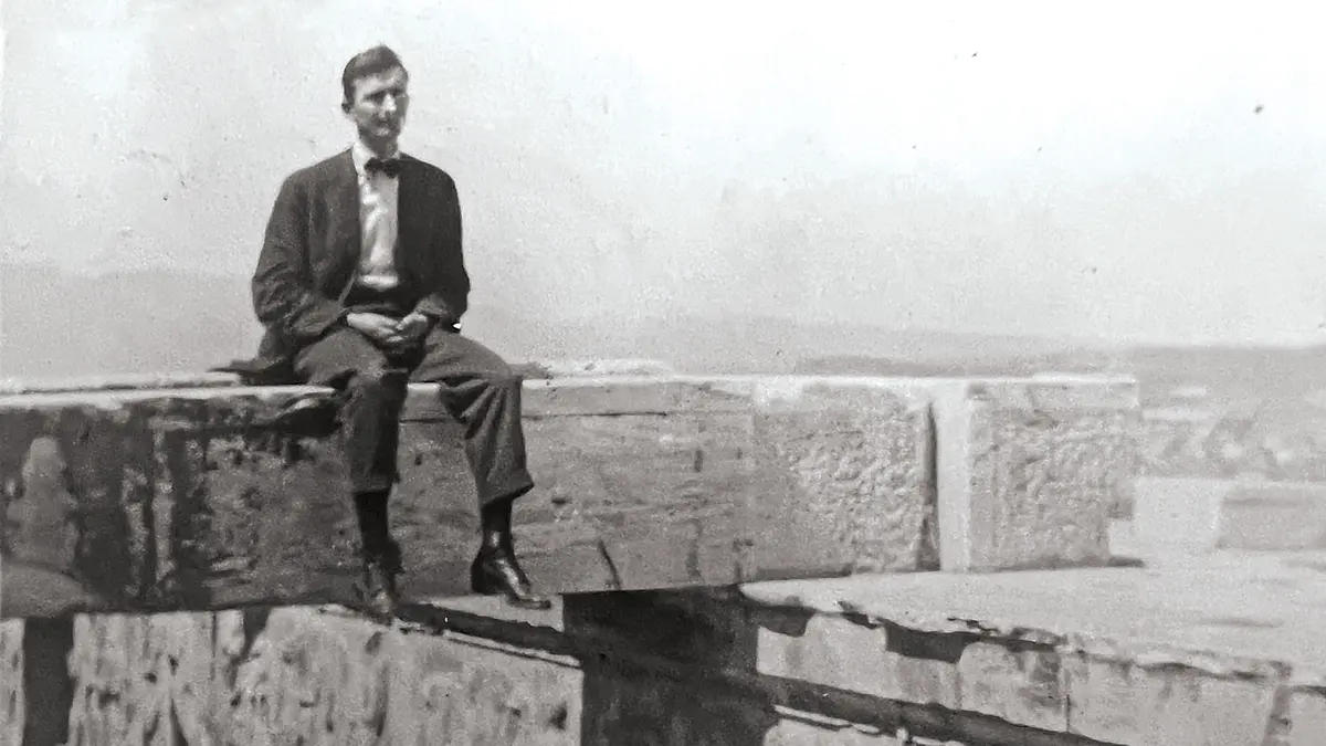 A man in a suit and bowtie sits on a slab of stone, which might be a fallen pillar, balanced on top of a stone floor at least 4 feet off the ground, which can’t be seen in this photo.