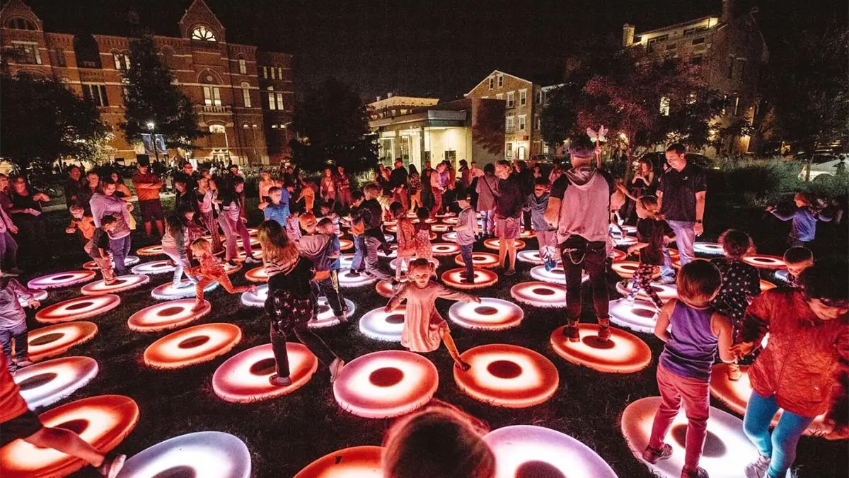 In the dark outside, a crowd of festival-goers including multiple kids play on lighted-up multicolored circles on the ground.