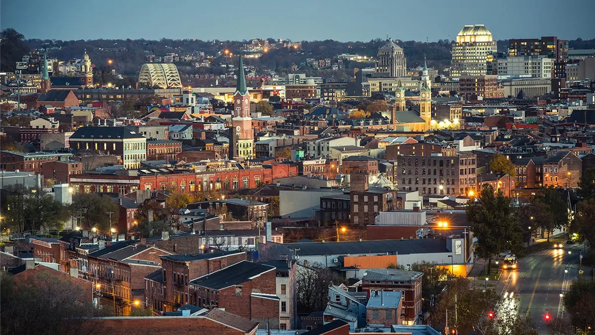 From a bird’s eye view as the sun goes down, lights come on in an area of Cincinnati that is mostly red brick buildings of a few stories. Here and there, taller ones stand out.