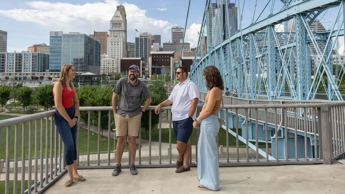 Four Buckeye alumni in their 30s, laughing and wearing summer clothes, make eye contact as they talk. In the distance are a blue-green bridge, part of the Cincinnati skyline and fluffy white clouds.